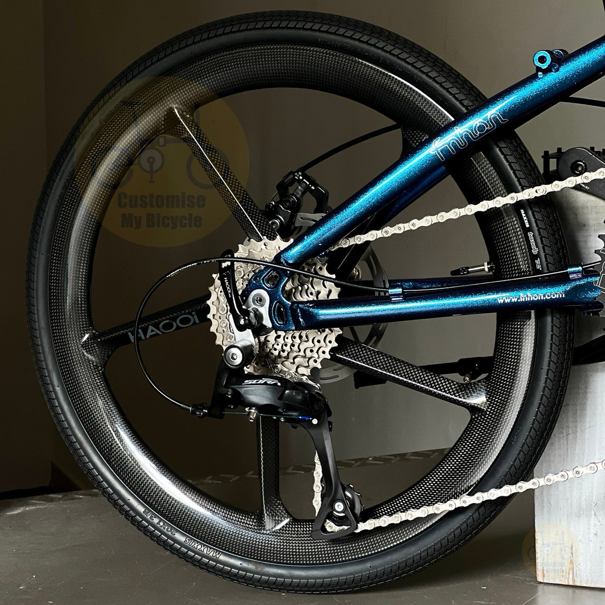 Close-up of a folding bike rear wheel and frame, showing a black carbon fiber wheel, silver cassette, and a metallic blue frame with 'HAOOi' branding.