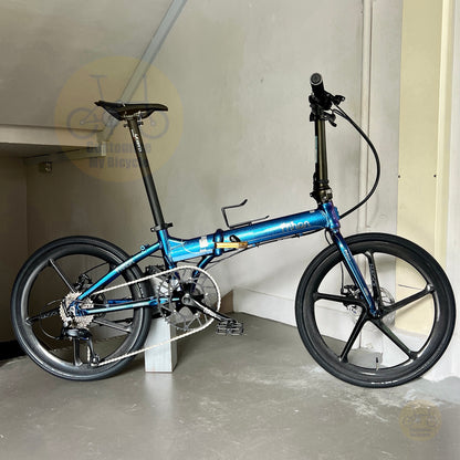 A blue and black folding bicycle with 20-inch wheels, shown in a garage setting with a concrete floor and beige walls.
