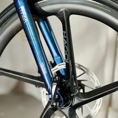 Close-up of a bicycle wheel and fork area showing a blue metallic frame with 'mion' branding, a carbon fiber wheel, and a white background.