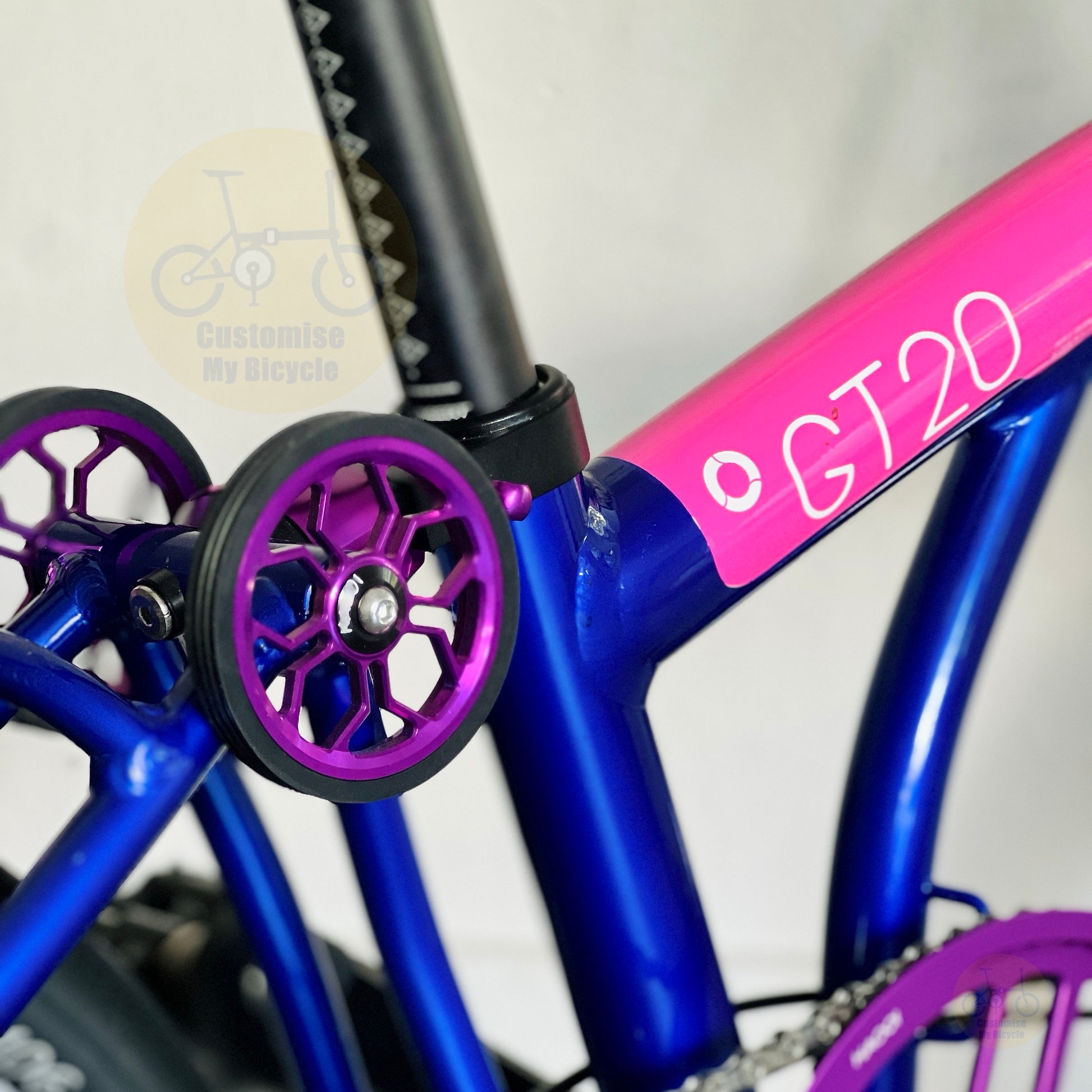 Close-up of a bicycle frame with bright blue tubing, a pink seat tube section labeled 'GT20', and purple training wheels with black tires. A faint 'Customise My Bicycle' watermark is visible in the upper left.