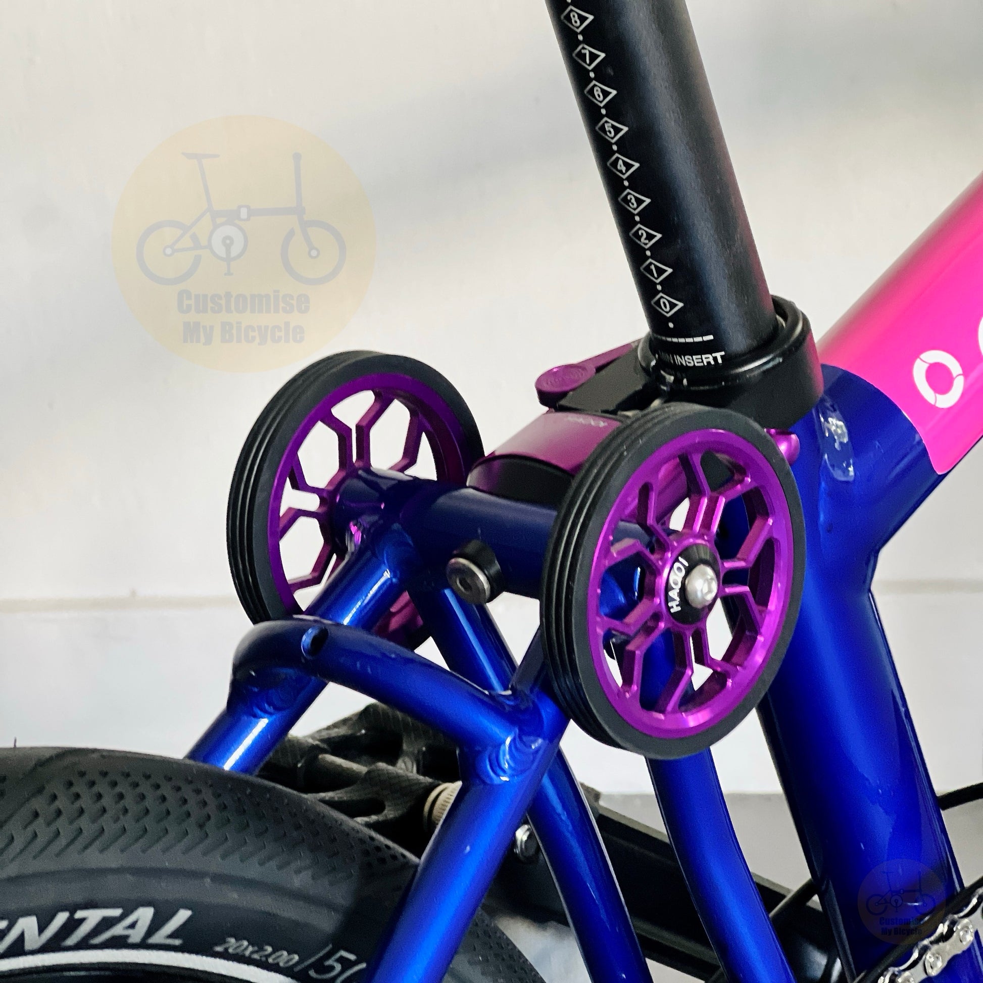 Close-up of a bicycle ezy wheel area showing purple and black wheel spokes, blue frame tubes, part of a pink and blue bike frame, and a visible 'Customize My Bicycle' watermark.