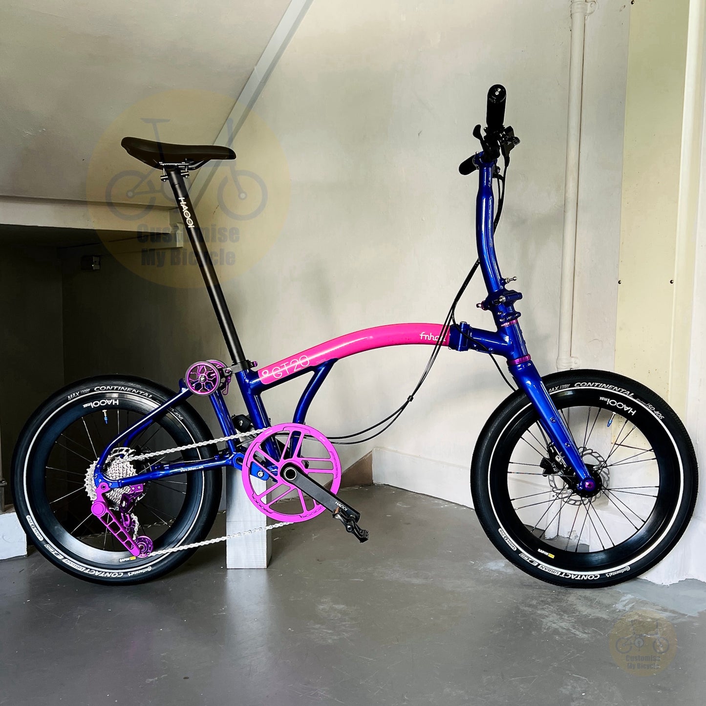 A folding bicycle with a vibrant blue frame and bright pink accents, including the seat tube, chainstays, and crankset, parked indoors against a beige wall.