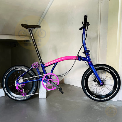 A folding bicycle with a vibrant blue frame and bright pink accents, including the seat tube, chainstays, and crankset, parked indoors against a beige wall.