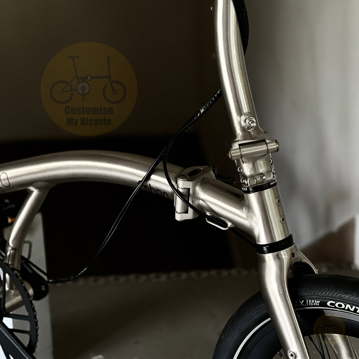 Close-up of a metallic silver folding bicycle frame with a black cable running through a clamp, set against a dark indoor background with a 'Customise My Bicycle' logo watermark.