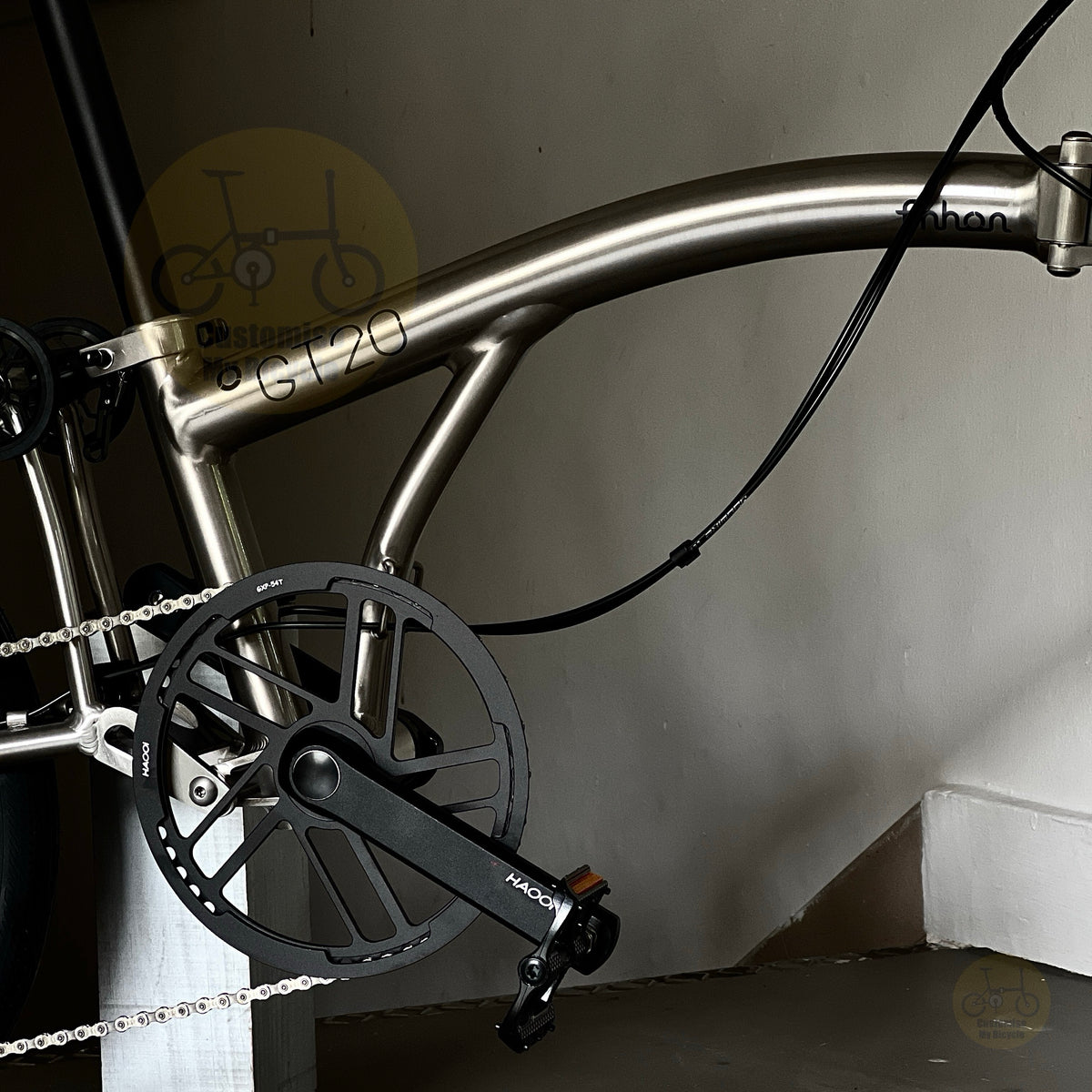 Close-up of a metallic silver folding bicycle frame with black crankset and chainring, photographed indoors against a plain wall.
