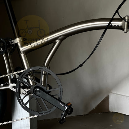 Close-up of a metallic silver folding bicycle frame with black crankset and chainring, photographed indoors against a plain wall.