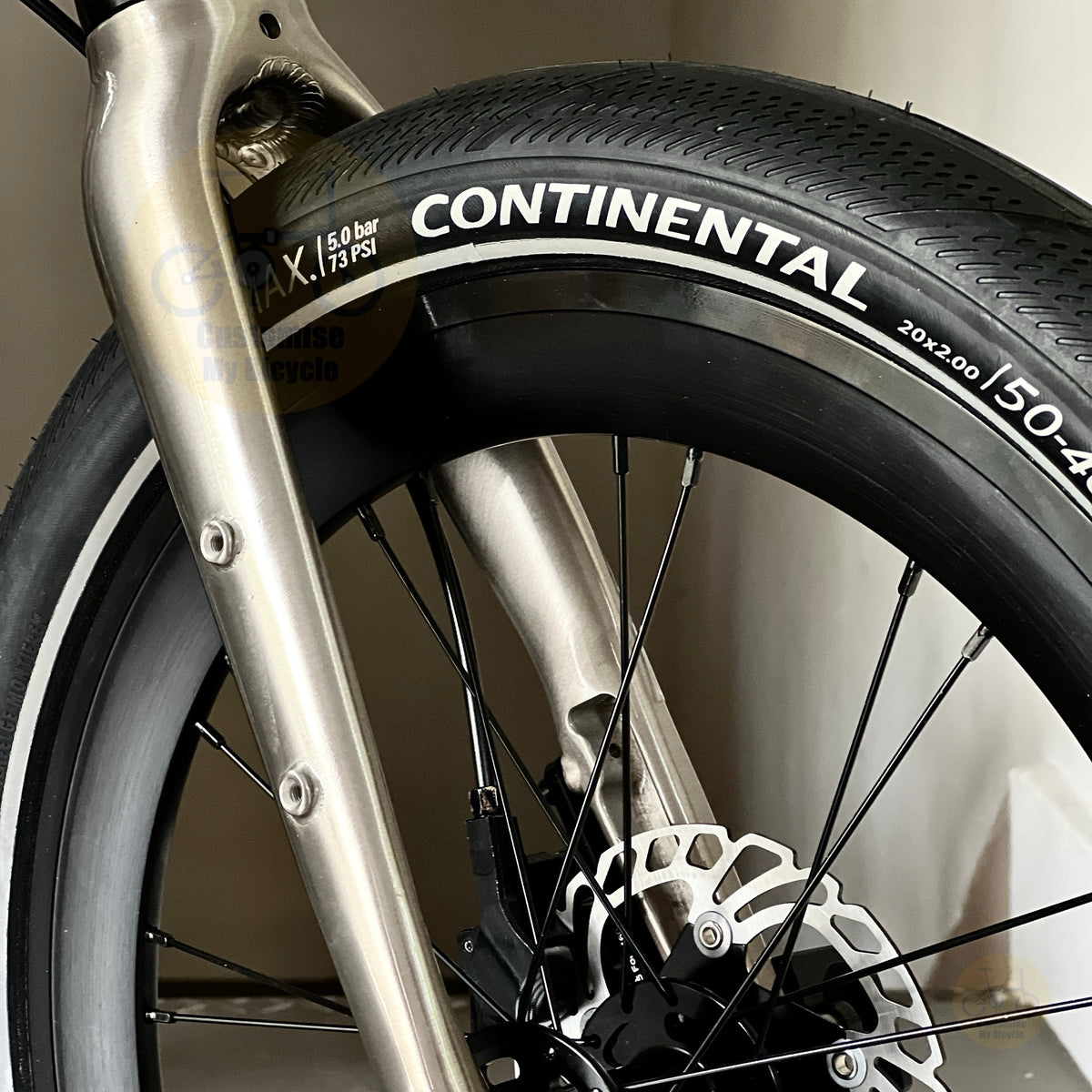 Close-up of a bicycle wheel with a Continental tire, silver front fork, and disc brake, photographed indoors against a light neutral wall.