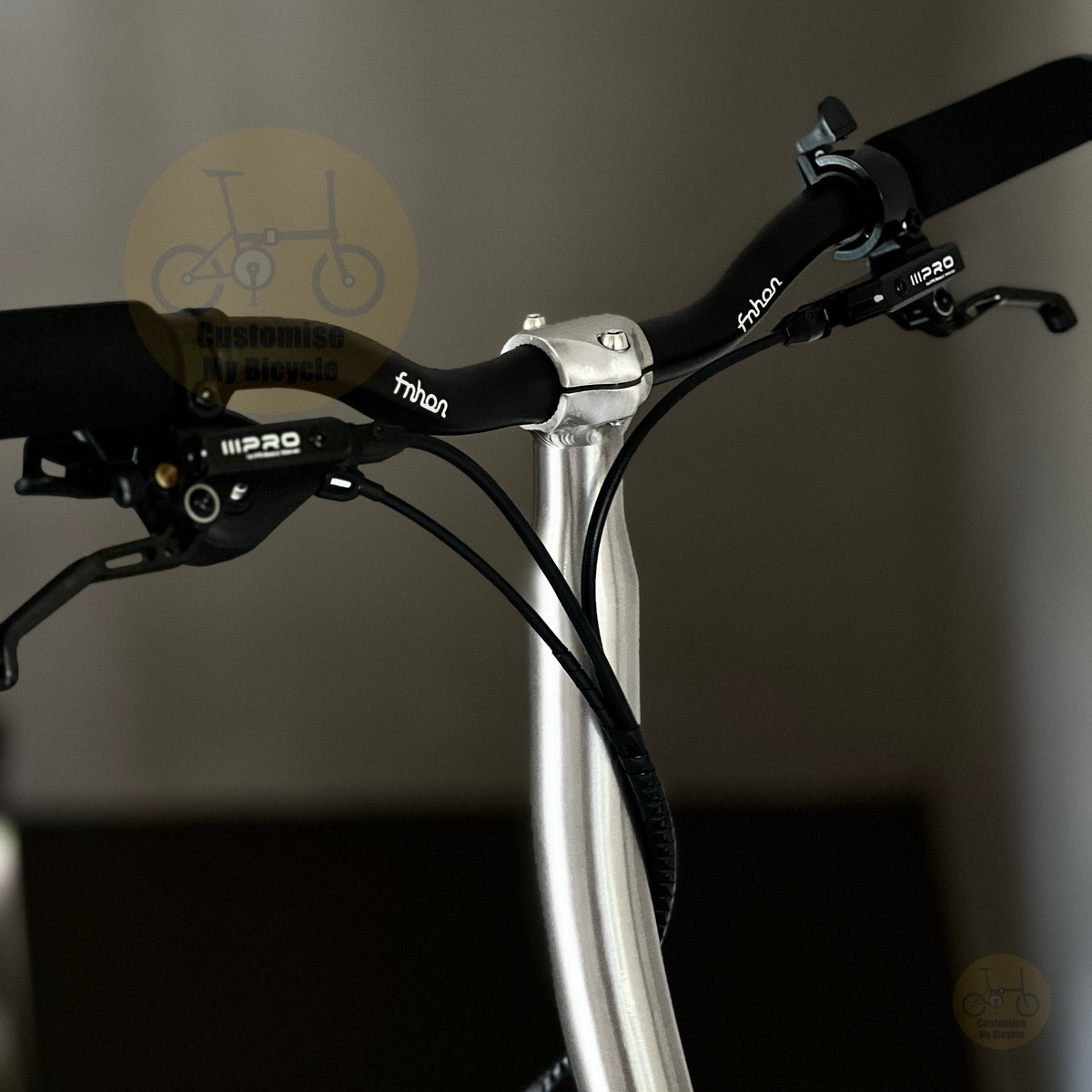 Close-up of a bicycle handlebar setup with black Frison brake levers and grips, a silver handlebar clamp, and black cables running down a silver stem against a dark gray background.