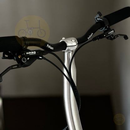 Close-up of a bicycle handlebar setup with black Frison brake levers and grips, a silver handlebar clamp, and black cables running down a silver stem against a dark gray background.