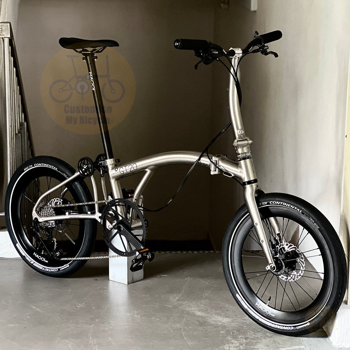 A silver metallic folding bicycle with black components, including tires, saddle, handlebars, and wheels, displayed indoors on a concrete floor against a beige wall.