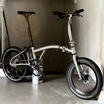 A silver metallic folding bicycle with black components, including tires, saddle, handlebars, and wheels, displayed indoors on a concrete floor against a beige wall.