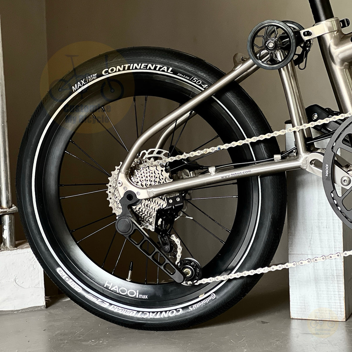 Close-up of a bicycle wheel with a Continental Maxxis 20" 406 tire, showing the tire sidewall branding, cassette, chainring, and part of the metallic titanium colour frame.
