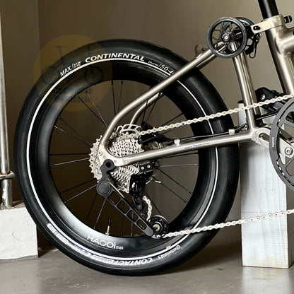 Close-up of a bicycle wheel with a Continental Maxxis 20" 406 tire, showing the tire sidewall branding, cassette, chainring, and part of the metallic titanium colour frame.