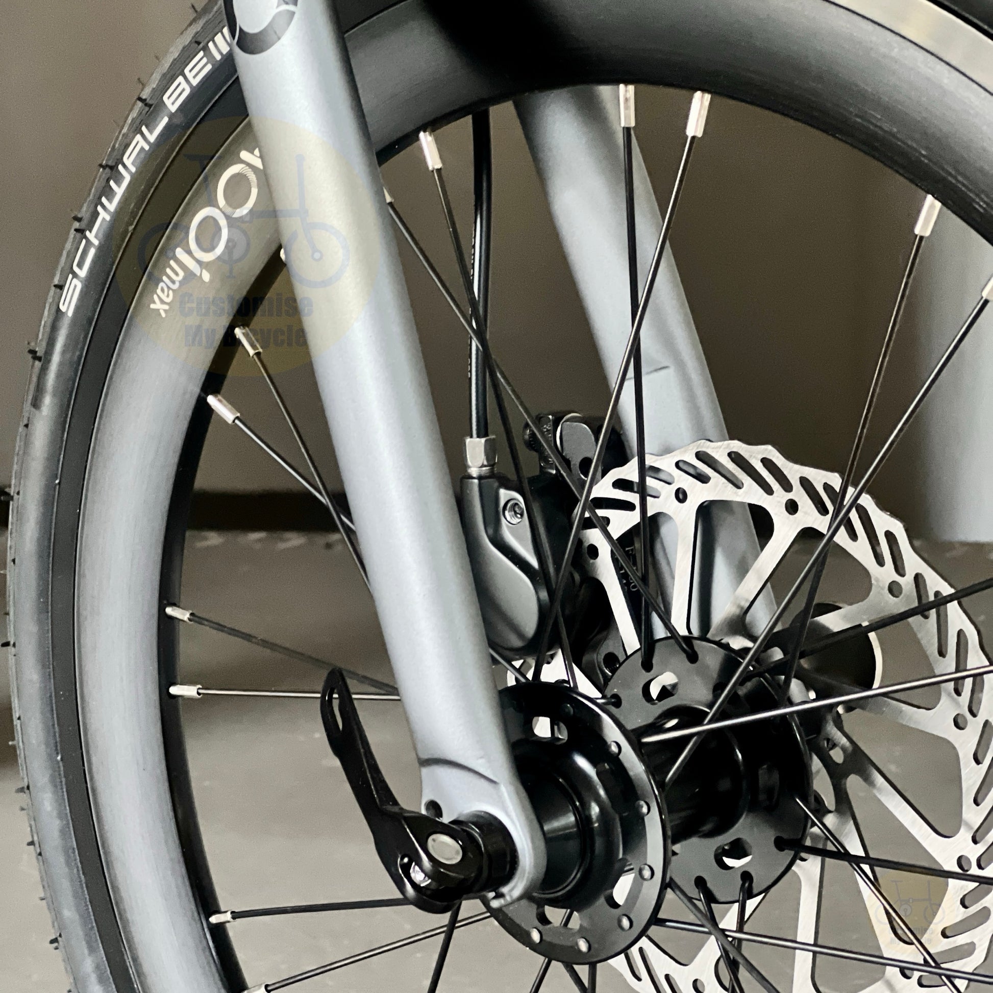 Close-up of a bicycle wheel and front fork showing a silver disc brake rotor, black spokes, and part of a black tire with white branding text.