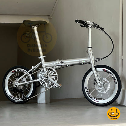 Lower side view of Fnhon Zephyr 18-inch folding bike in polished silver, displayed on wooden deck with city skyline at sunset.
