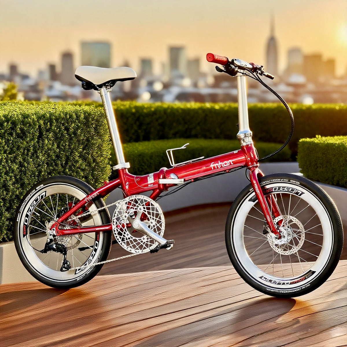 A red folding bicycle with white wheels and silver components, parked on a wooden deck with a city skyline and green hedges in the background.